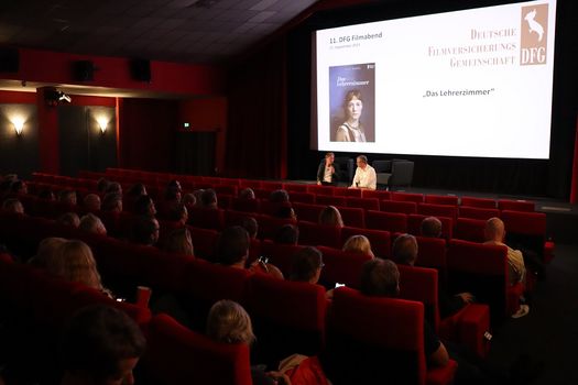 Kinoleinwand mit dem Filmplakat von "Das Lehrerzimmer" und dem Text "11. DFG-Filmabend". Davor ein Kinopublikum auf roten Plüschsesseln